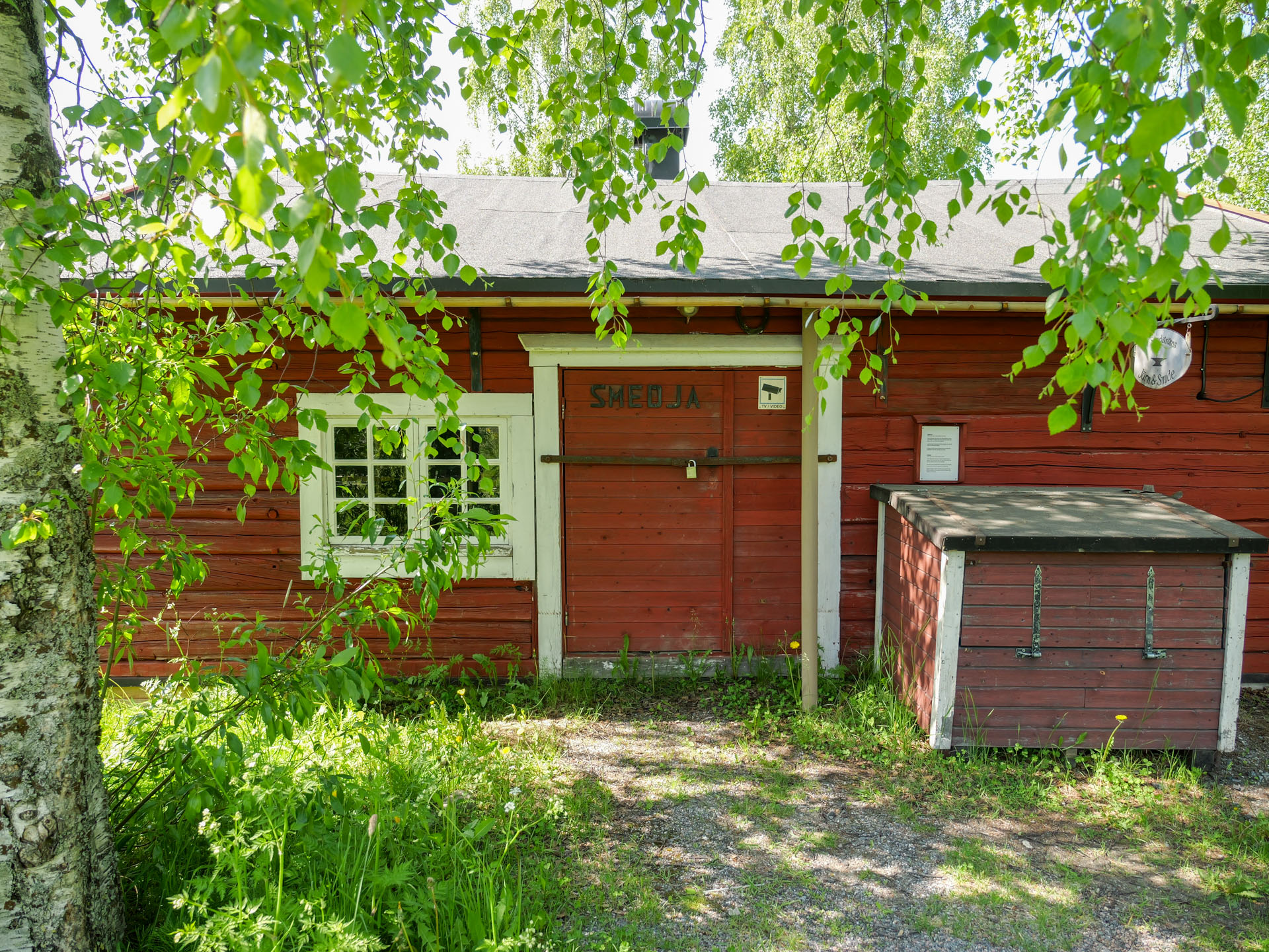 Smedjan från utsidan. En röd träbyggnad omgiven av grönskande träd